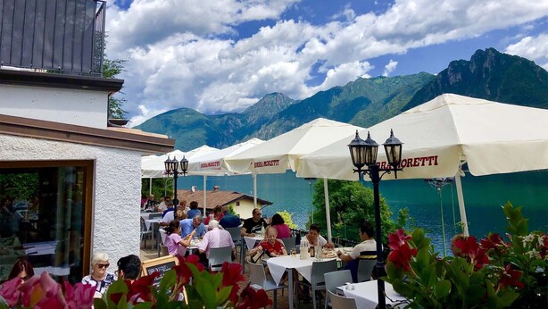 An outdoor dining area with tables and umbrellas, overlooking a lake and mountains. - Directly on the lake (Crone)