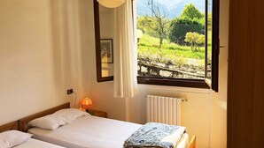 A bedroom with a bed, a bedside table with a lamp, a window with a view of greenery and a fence, and a radiator. - Directly on the lake (Crone)