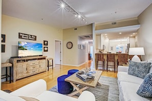 A modern living room with a television, a coffee table, and a sofa. There is a kitchen area in the background with a bar and seating.