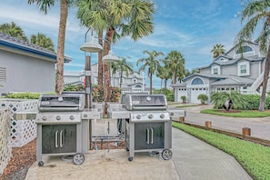 A patio area with two barbecue grills, a white fence, and a street view with palm trees and houses.