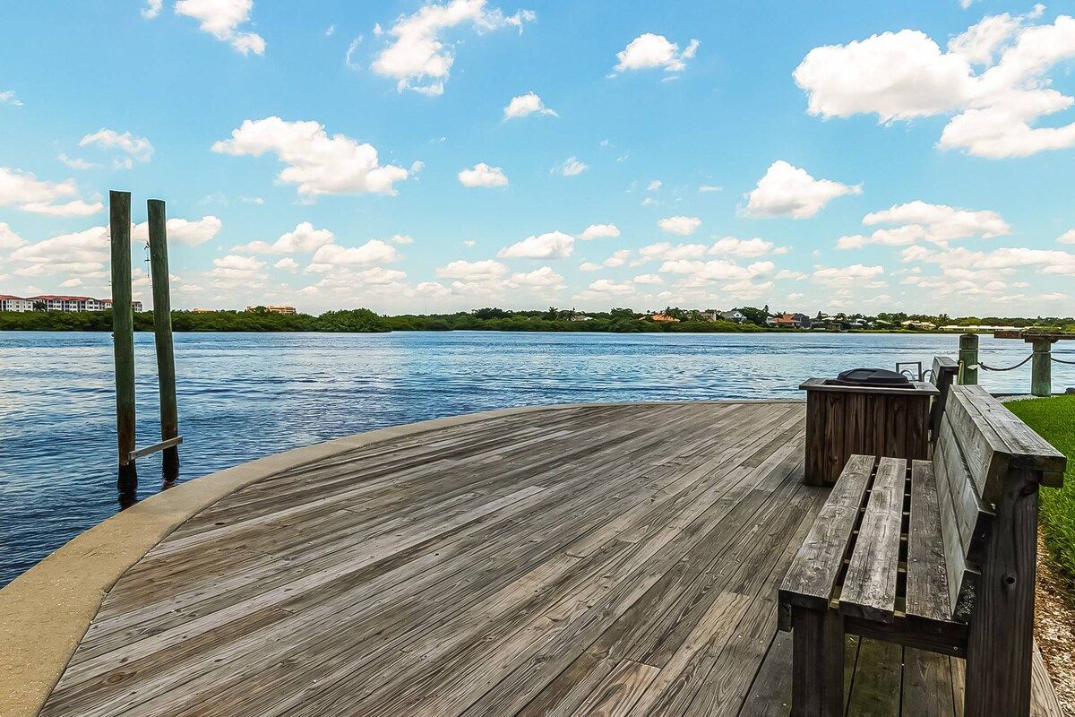 A wooden deck with a bench and a table, overlooking a body of water with a distant shoreline.