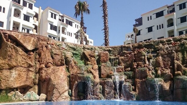 A pool with clear blue water, surrounded by rocky terrain and palm trees. - Beautiful apartment in Lilacs Garden 4539 (Cabo Negro)