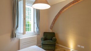 Un dormitorio con un sillón verde, una ventana con cortinas, un radiador blanco y un arco de ladrillo.