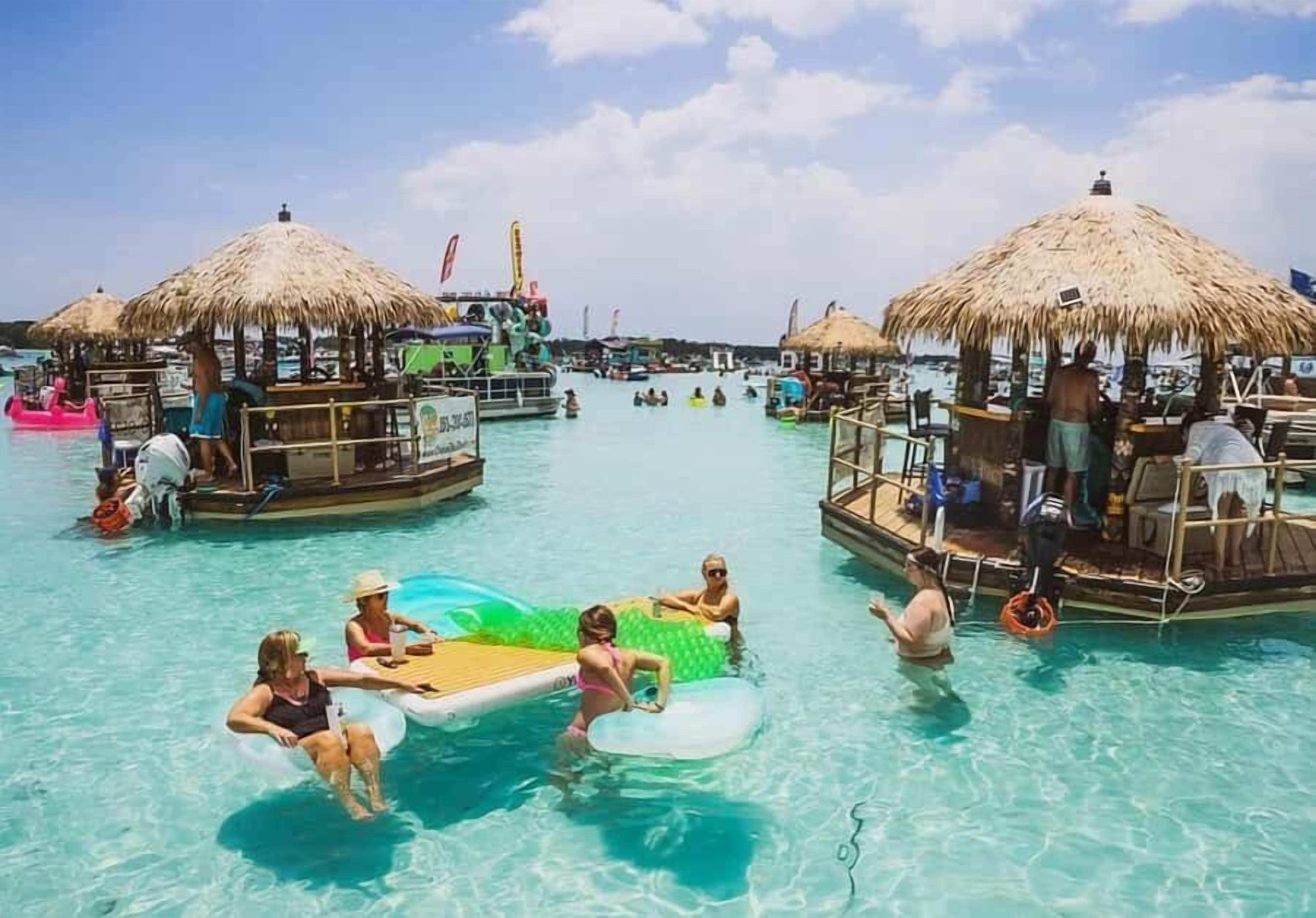 A tropical resort with clear blue water, thatched-roof cabanas, and a floating bar area.
