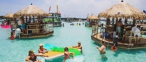 Un resort tropical con aguas azul cristalino, cabañas de techo de paja y un área de bar flotante.