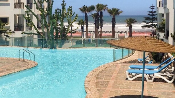 A pool with clear blue water, surrounded by lounge chairs and a thatched umbrella. - Sublime beachfront apartment 4317 (Dar Bouazza)