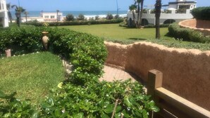 A landscaped garden with a view of the sea, a palm tree, and a building with a tower. - Sublime beachfront apartment 4317 (Dar Bouazza)