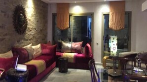 A living room with a red sofa, a coffee table, and a TV. There is a stone wall and a window with curtains. - Sublime beachfront apartment 4317 (Dar Bouazza)