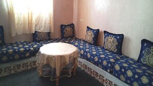 Una stanza con un tavolo rotondo ricoperto da una tovaglia all'uncinetto, un divano con cuscini decorati e una finestra con tende leggere.