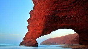 Un'imponente formazione rocciosa ad arco naturale su una spiaggia, con acque calme e colline in lontananza.