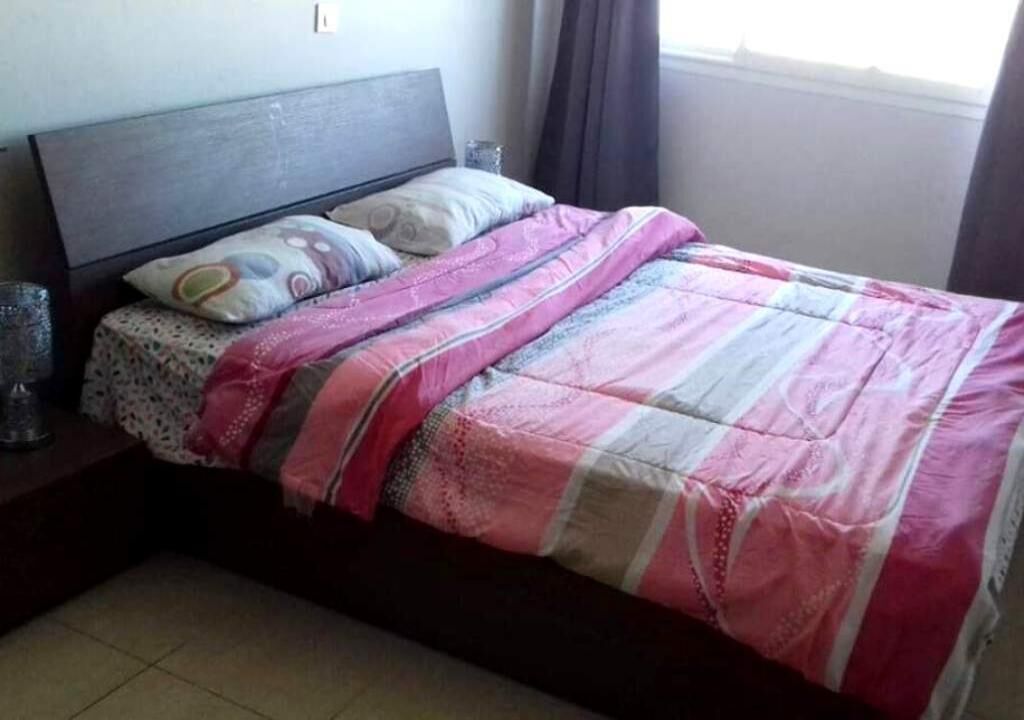 Una cama con colcha estampada y almohadas, una mesita de noche con un vaso, y una ventana con cortinas.