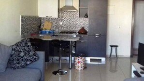 A compact kitchen with a gas stove, a microwave, and a built-in oven. There is a grey sofa with a patterned pillow and a small stool.