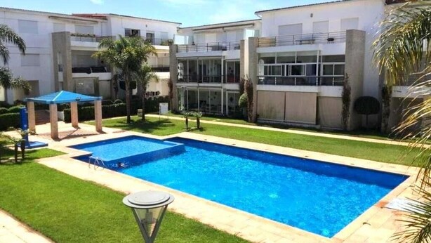 A swimming pool surrounded by a well-maintained lawn, with a building featuring balconies and large windows in the background. - Residence The Pearls of Tamaris 4457 (Tamaris)