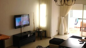 A living room with a flat-screen TV, a dark wooden entertainment unit, a small table with a lamp, and a large window with curtains. - Sublime apartment with sea and golf views 4301 (Asilah)