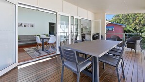 Un área de comedor moderna al aire libre, con una mesa grande y sillas, conectada a una puerta de cristal que da a una sala de estar con sofá y sillones.
