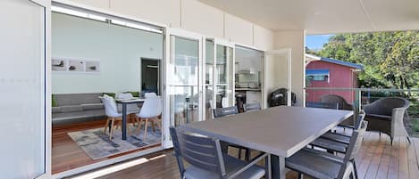 Un espace repas extérieur moderne, doté d’une grande table et de chaises, et relié à un salon par une porte vitrée, ce salon comprenant un canapé et des fauteuils.