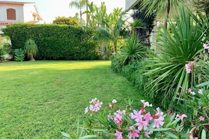 Um jardim bem cuidado com gramado, diversas plantas e um edifício ao fundo.