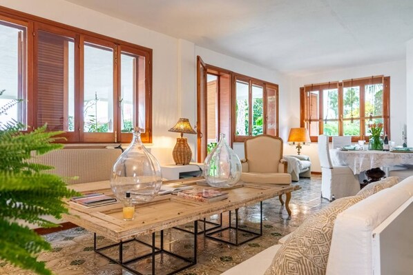 A room with a wooden coffee table, two lamps, a sofa, and large windows with wooden shutters.