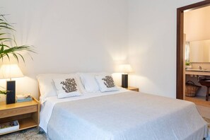 Uma cama bem arrumada com roupa de cama branca e almofadas decorativas, duas luminárias de cabeceira, uma mesinha de cabeceira com uma revista e um vaso com planta.