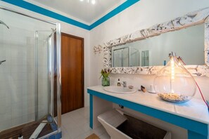 A bathroom with a glass shower enclosure, a sink with a marble countertop, and a mirror with a decorative frame.