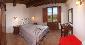 A bedroom with a bed, a desk, a chair, and a window with a view of the countryside. - Ortensia Apartment 4 + 4 (Lajatico)