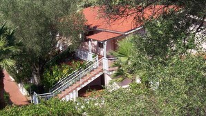 Une maison avec un toit rouge, un escalier menant à un balcon, le tout entouré d’une végétation luxuriante.