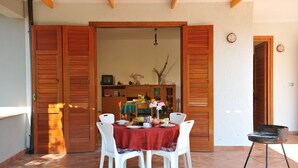 Un coin repas avec une table ronde, des chaises blanches et une nappe rouge. On y trouve une porte en bois avec des volets, un petit téléviseur posé sur un meuble et un vase rempli de fleurs.
