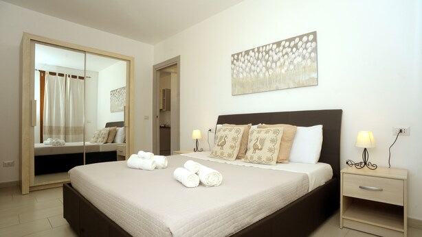 A neatly made bed with rolled towels, a bedside table with a lamp, and a large mirror. - ALKIRA LODGE - ILUKA Two-bedroom apartment with sea view (Alghero)