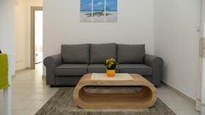 A living room with a grey sofa, a wooden coffee table with a plant, and a beach-themed wall art. - ALKIRA LODGE - ILUKA Two-bedroom apartment with sea view (Alghero)