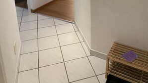 Un couloir au sol carrelé de carreaux blancs, une porte en bois au fond et un miroir rond accroché au mur.