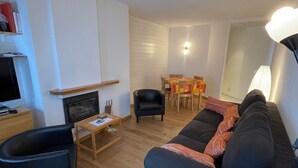 Un salon moderne avec un canapé noir, une table basse en bois et un téléviseur posé sur un meuble. On aperçoit à l’arrière-plan un coin repas avec une table et des chaises.
