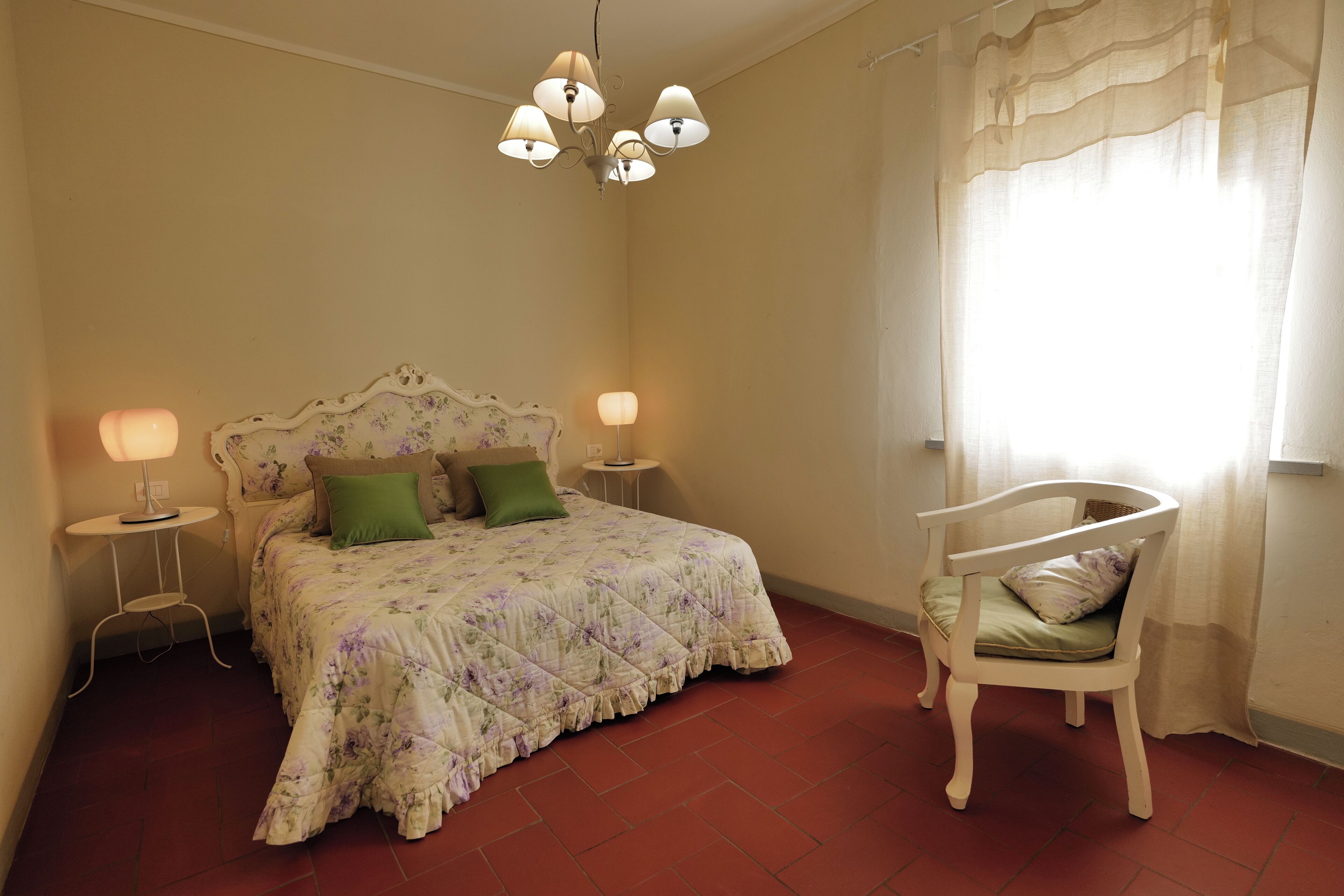 Une chambre à coucher avec une couvre-lit à motifs floraux, deux lampes de chevet, un coussin vert, une chaise blanche et un lustre.