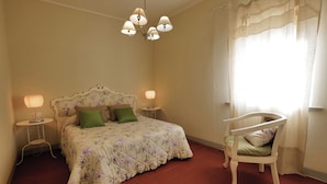 A bedroom with a floral bedspread, two bedside lamps, a green pillow, a white chair, and a chandelier. - AGRITURISMO IL PRINCIPINO Apartment 6 (San Donato)