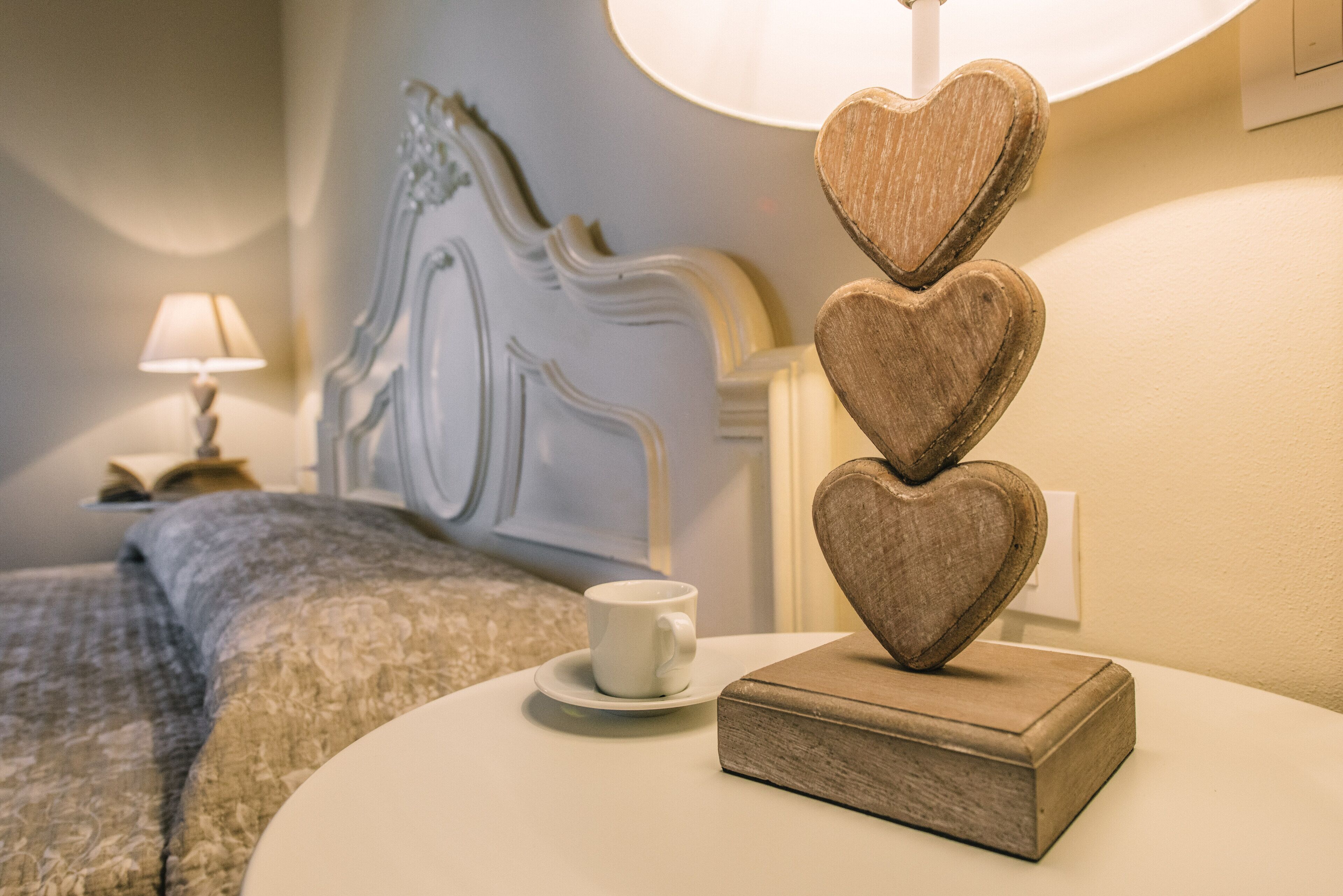 Habitación de hotel ordenada con cama, mesita de noche con lámpara, taza y un objeto decorativo con forma de corazón.
