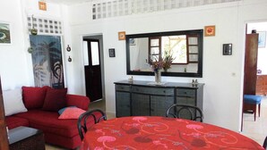 Un coin repas avec une table ronde recouverte d’une nappe rouge, un canapé rouge et une chaise en métal noir. On y trouve également un meuble surmonté d’un miroir et un vase avec des fleurs placé sur la table.