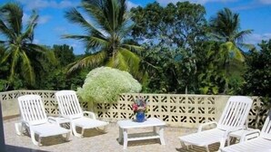 Une terrasse dotée de chaises longues blanches et d’une petite table ornée d’un vase de fleurs, le tout entouré d’une clôture en treillis de bois et de palmiers.