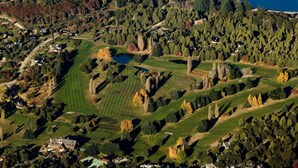 An aerial view of a golf course with multiple greens, a lake, and surrounding wooded areas.