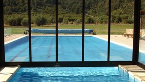 An indoor swimming pool with clear blue water, surrounded by a covered area and a view of a forested hillside.