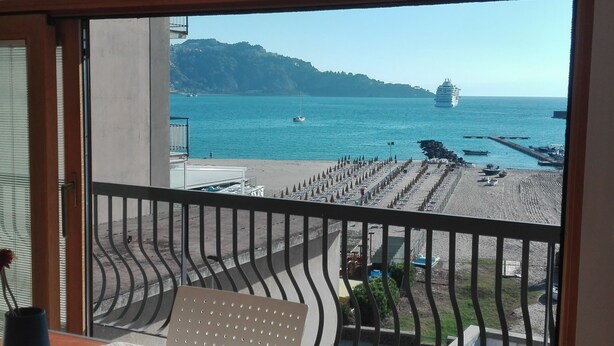 A balcony with a view of a beach, a yacht, and a distant mountain.