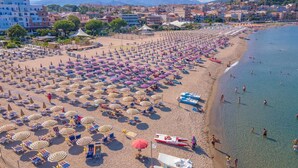 A beach with numerous umbrellas and sun loungers, a lifeguard boat, and a clear view of the sea and distant mountains.