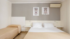 A hotel room with a bed, a bedside table, a small desk, and two wall-mounted air conditioning units.