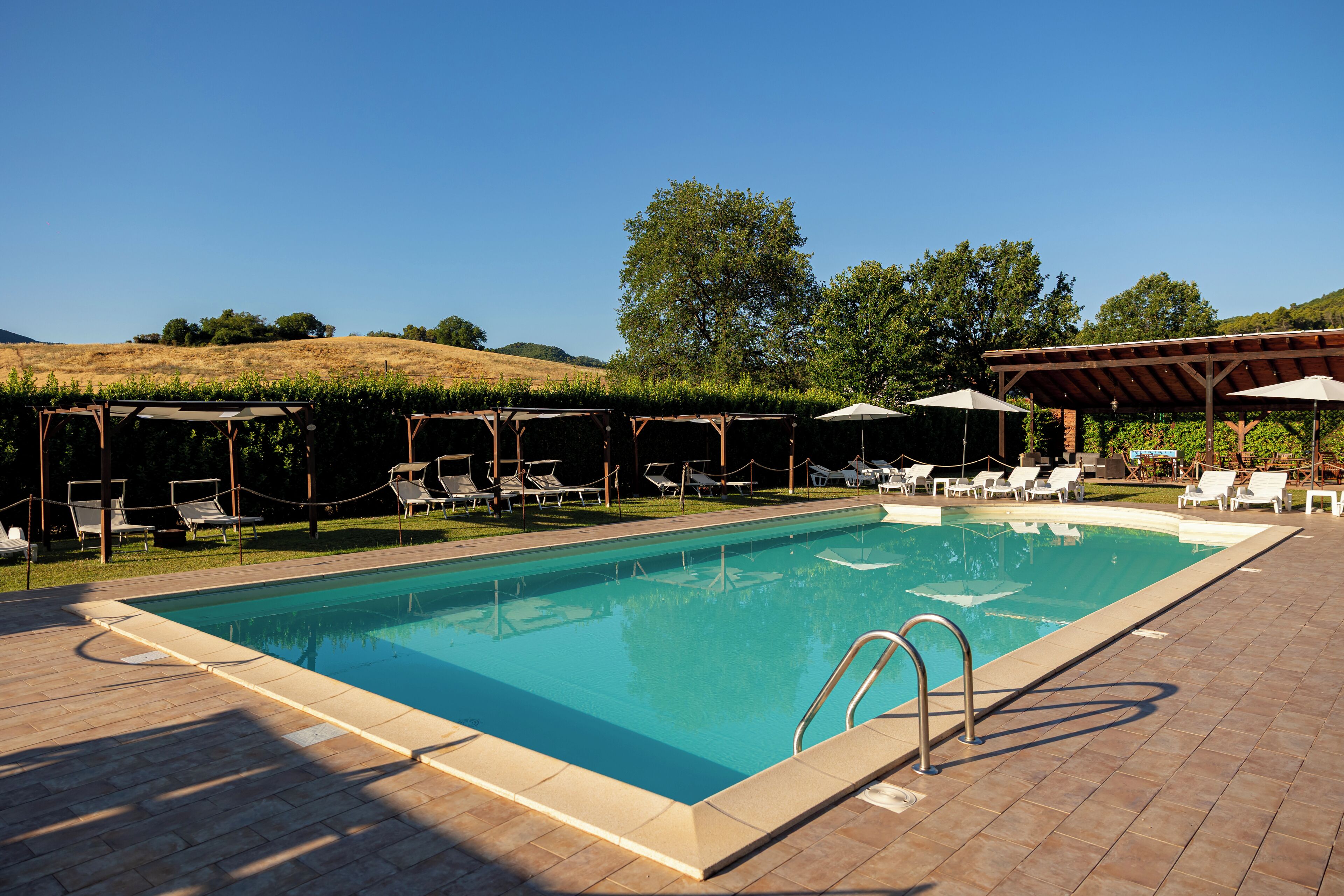 Una piscina rectangular con un borde empedrado, rodeada de sillas de descanso y sombrillas.