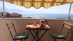 Ein Tisch mit zwei Stühlen unter einem gestreiften Sonnendach mit Blick auf eine malerische Landschaft aus Meer und Bergen.