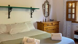 Ein ordentlich bezogenes Bett mit Kissen und gefalteten Handtüchern, eine hölzerne Kommode mit Schubladen, ein Spiegel und ein Fenster mit Vorhängen.