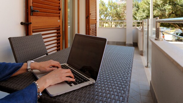 A person using a laptop on a balcony with a view of trees and buildings. - ALKIRA LODGE - Residential Hotel Apartments (Alghero)