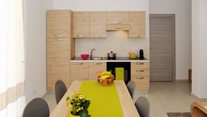 A modern kitchen with wooden cabinets, a dining table with chairs, and a fruit basket. - ALKIRA LODGE - Residential Hotel Apartments (Alghero)