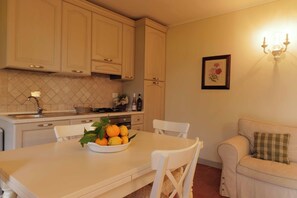 Una cocina con armarios blancos, una mesa de comedor con sillas, un cuenco de naranjas y un sofá con un cojín de cuadros.