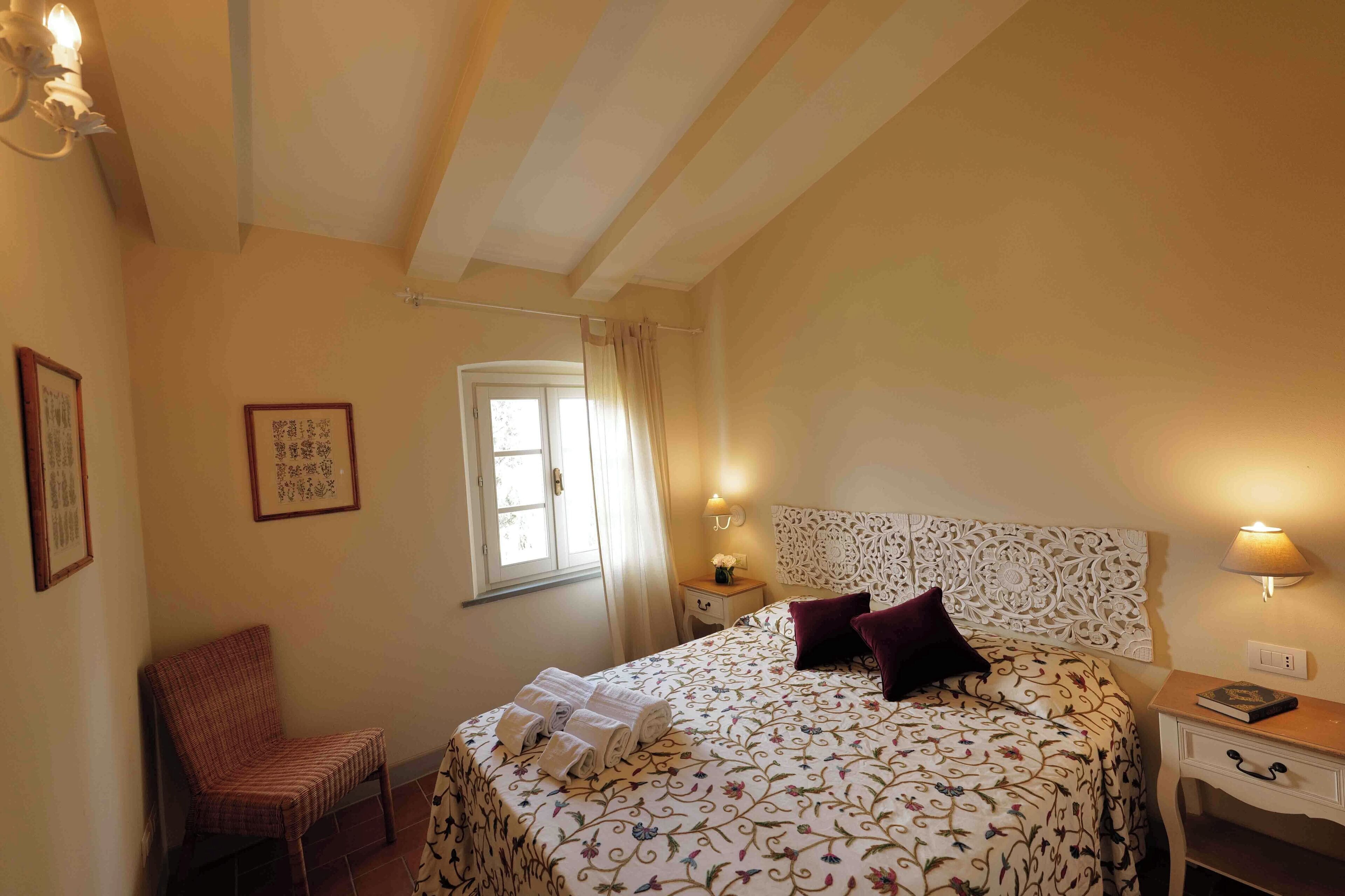 Ein Schlafzimmer mit Bett, Stuhl, Nachttisch, Lampe und einem Fenster mit Vorhängen.