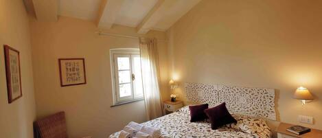 A bedroom with a bed, a chair, a nightstand, a lamp, and a window with curtains.
