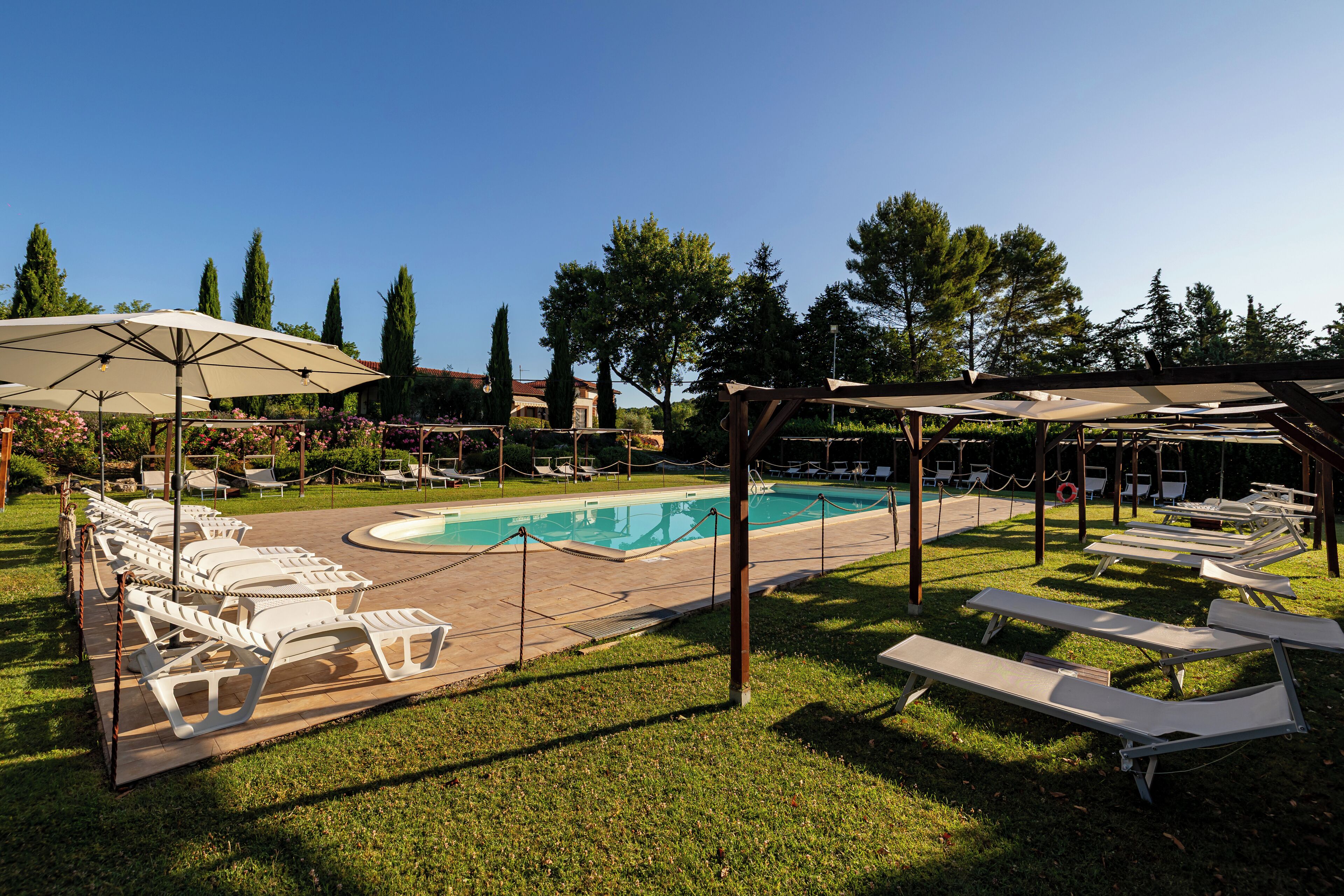A poolside area with lounge chairs and umbrellas, surrounded by trees and a clear sky.